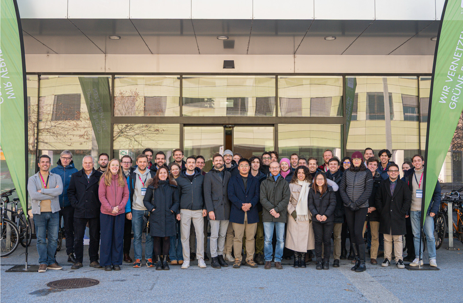 A large group of people smiles while posing in front of a modern building between two green flags reading ENERGIENETZE STEIERMARK – WIR VERNETZEN GRÜNE ENERGIE!