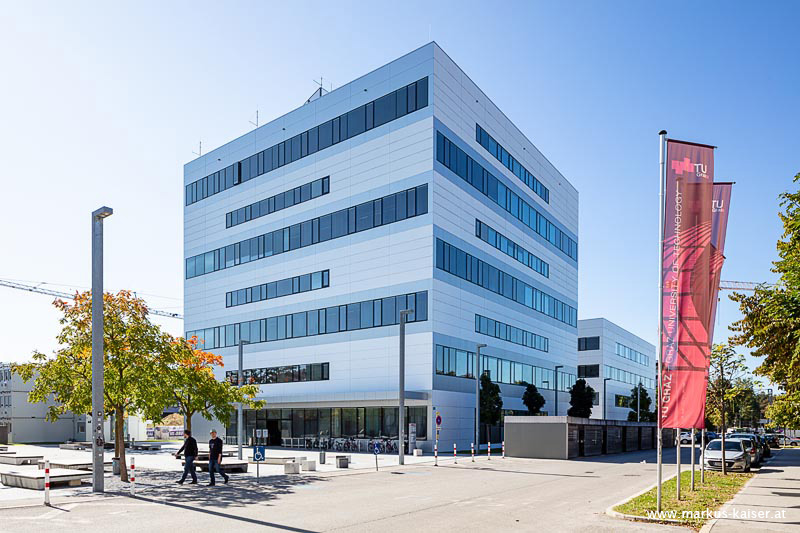 Modern multi-story TU Graz building with large glass windows, flanked by trees, streetlights, two walking people, and red TU flags in the foreground.
